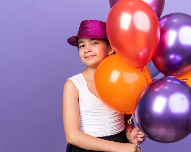 Довольная молодая кавказская девушка в фиолетовой шляпе держит гелиевые шары, изолированные на фиолетовой стене с копией пространства