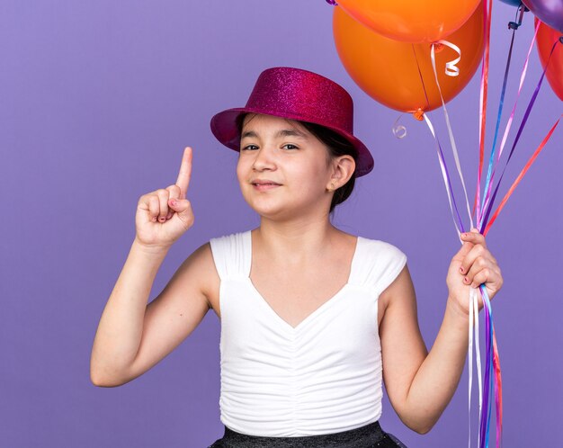 pleased young caucasian girl with violet party hat holding helium balloons and pointing up isolated on purple wall with copy space