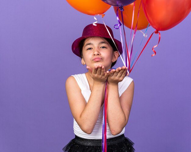 pleased young caucasian girl with violet party hat holding helium balloons and sending kiss with hands isolated on purple wall with copy space