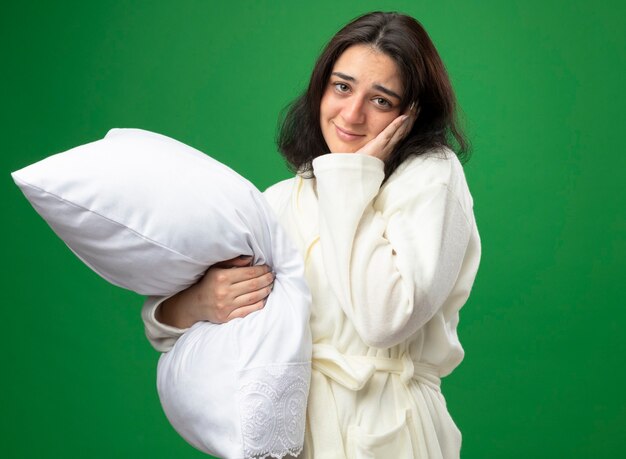 Pleased young caucasian ill girl wearing robe holding pillow keeping hand on face looking at camera isolated on green background