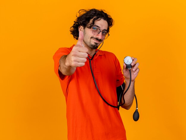 コピースペースでオレンジ色の背景に分離された親指を示すカメラを見て血圧計を保持している眼鏡と聴診器を身に着けている若い白人の病気の人を喜ばせる