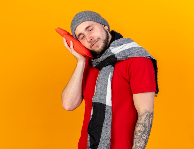 Pleased young caucasian ill man wearing winter hat and scarf puts head on hot water bottle on orange