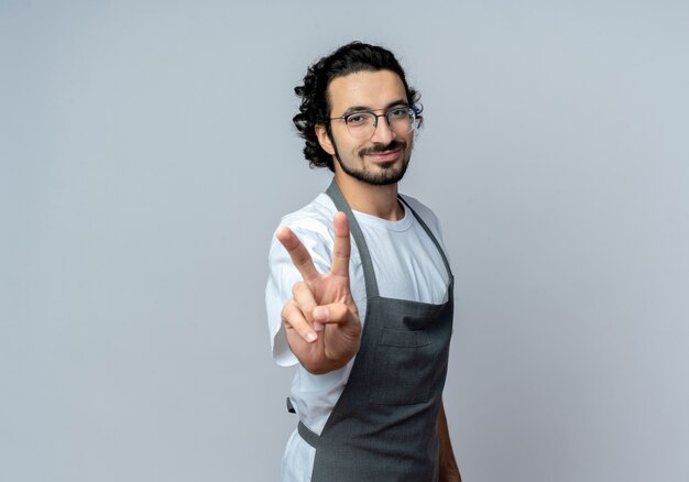 Pleased young caucasian male barber wearing glasses and wavy hair band in uniform doing peace sign isolated on white background with copy space