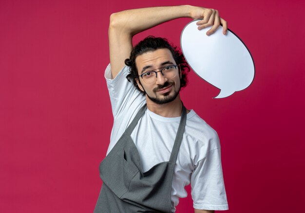 Pleased young caucasian male barber wearing glasses and wavy hair band in uniform holding chat bubble isolated on crimson background with copy space