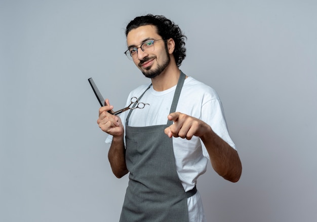 Pleased young caucasian male barber wearing glasses and wavy hair band in uniform holding comb and scissors pointing at camera isolated on white background with copy space