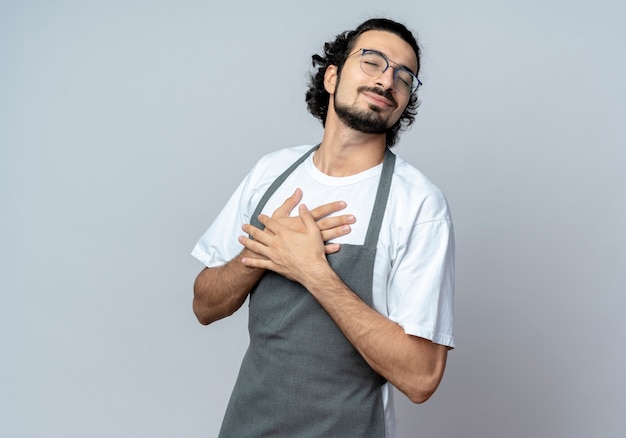 Pleased young caucasian male barber wearing glasses and wavy hair band in uniform putting hands on chest with closed eyes isolated on white background with copy space