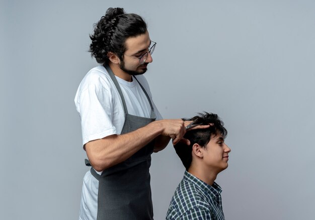 Pleased young caucasian male barber wearing glasses and wavy hair band in uniform standing in profile view doing haircut for his young client