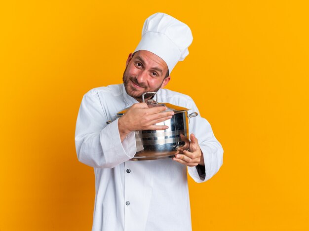 Pleased young caucasian male cook in chef uniform and cap holding and hugging pot 