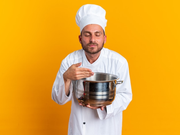 Felice giovane maschio caucasico cuoco in uniforme da chef e cappello che tiene la pentola tenendo la mano su di esso annusando con gli occhi chiusi