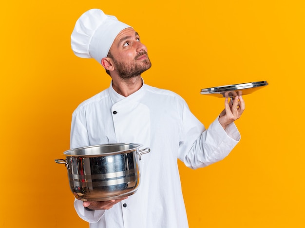 Free photo pleased young caucasian male cook in chef uniform and cap holding pot and pot lid looking up