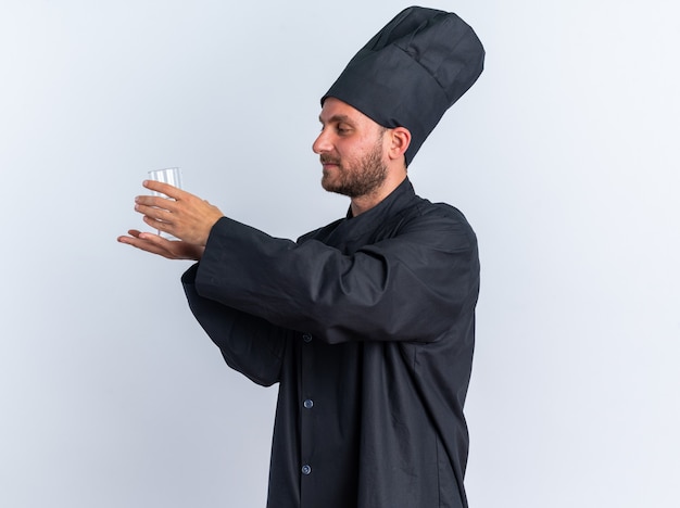 Pleased young caucasian male cook in chef uniform and cap standing in profile view trying to hold glass of water in his palm looking at it isolated on white wall