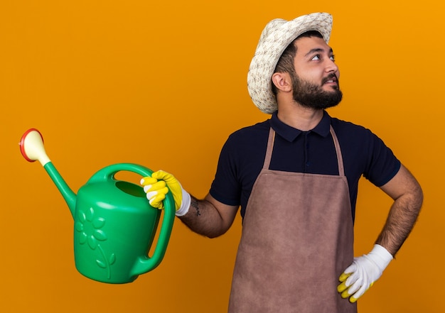Pleased young caucasian male gardener wearing gardening hat and gloves holding watering can looking at side isolated on orange wall with copy space