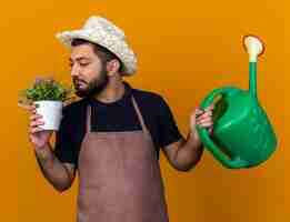 Free photo pleased young caucasian male gardener wearing gardening hat holding watering can and sniffing flowers in flowerpot isolated on orange wall with copy space