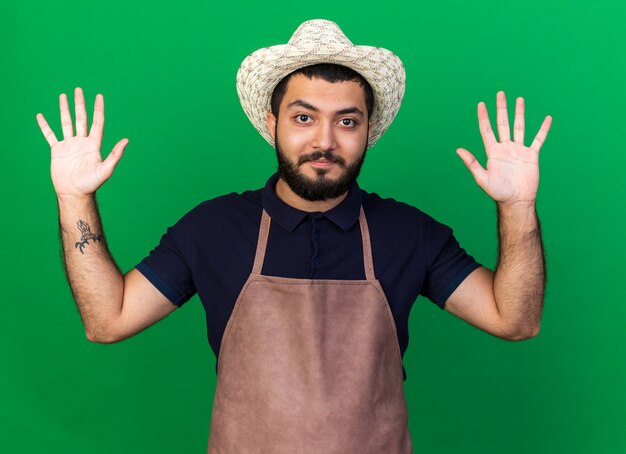 pleased young caucasian male gardener wearing gardening hat standing with raised hands isolated on green wall with copy space