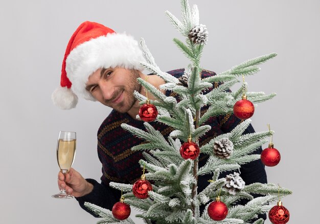 pleased young caucasian man wearing christmas hat standing behind christmas tree looking  holding glass of champagne winking isolated on white wall