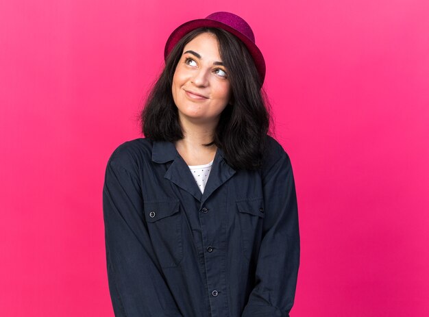 Pleased young caucasian party woman wearing party hat looking up isolated on pink wall