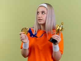 Free photo pleased young caucasian sporty girl with braces wearing headband and wristbands holds gold medal and winner cup looking at side