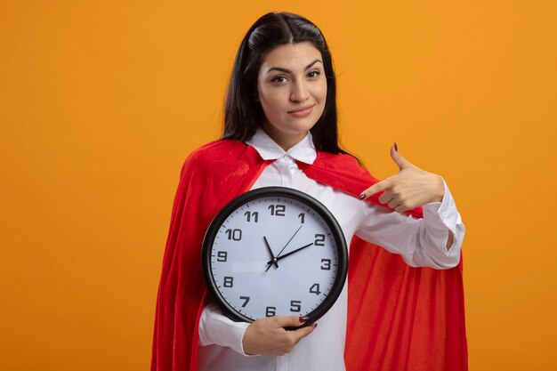 Pleased young caucasian superhero girl holding and pointing at clock looking at camera isolated on orange background with copy space