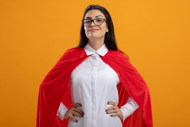 Free photo pleased young caucasian superhero girl wearing glasses keeping hands on waist looking at camera isolated on orange background