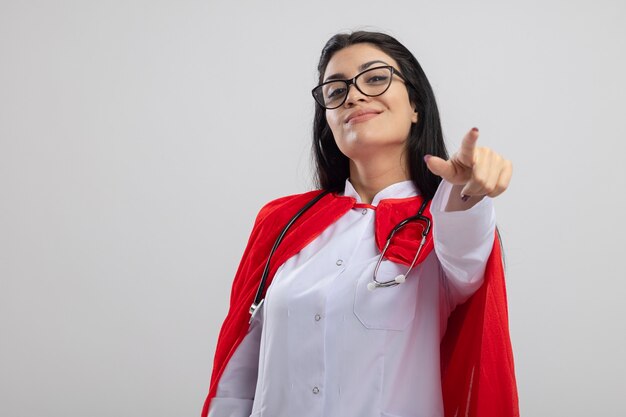 Pleased young caucasian superhero girl wearing glasses and stethoscope looking and pointing at camera isolated on white background with copy space