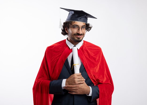 Довольный молодой кавказский супергерой в оптических очках в костюме с красным плащом и выпускной кепкой держит диплом на белом фоне с копией пространства