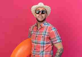 Free photo pleased young caucasian traveler man with straw beach hat in sun glasses stucks out tongue holding swim ring isolated on pink wall with copy space