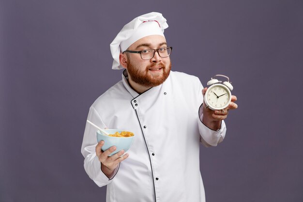 Pleased young chef wearing glasses uniform and cap holding bowl of macaroni pasta with spoon in it looking at camera showing alarm clock isolated on purple background