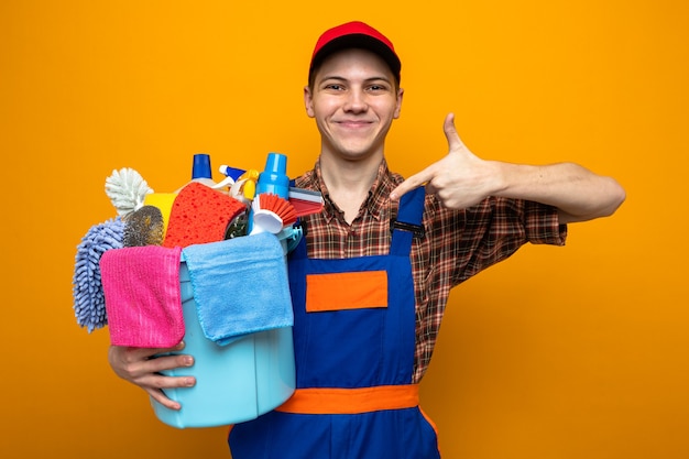 Довольный молодой уборщик в униформе и кепке держит и указывает на ведро с чистящими инструментами