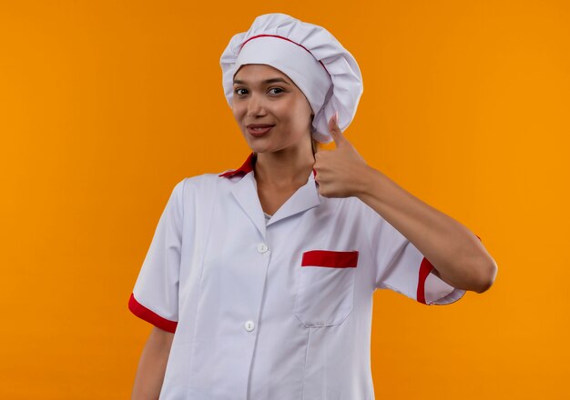 孤立したオレンジ色の壁に彼女の親指を制服シェフを着て喜んで若い料理人の女性