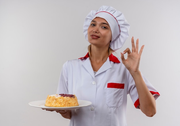 Pleased young cook female wearing chef uniform holding cake on plate showing okey gesture on isolated white wall with copy space