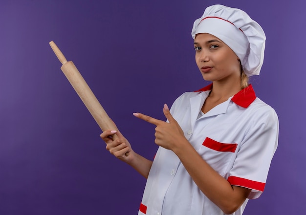 Довольная молодая женщина-повар в униформе шеф-повара указывает пальцем на скалку в руке на изолированной стене