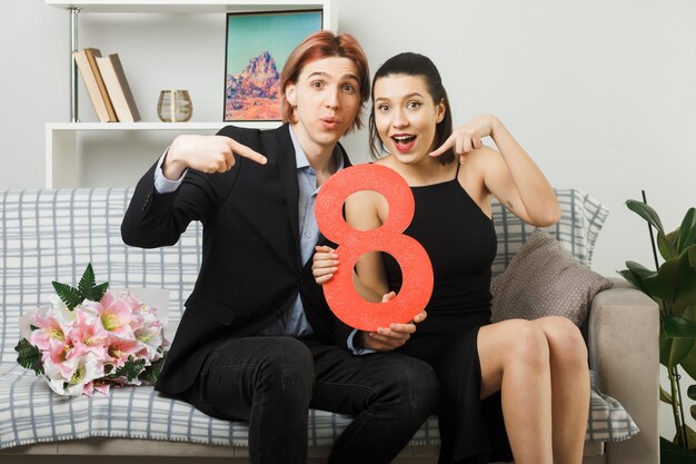 Довольная молодая пара в день счастливой женщины держит и указывает на номер восемь, сидя на диване в гостиной