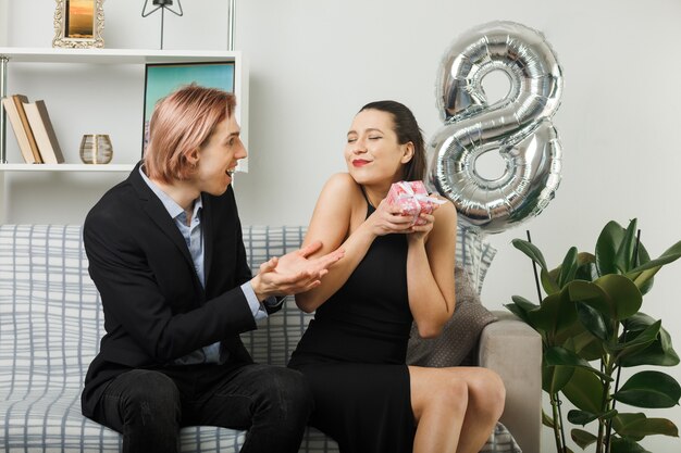 Довольная молодая пара в счастливый женский день держит подарок, сидя на диване в гостиной