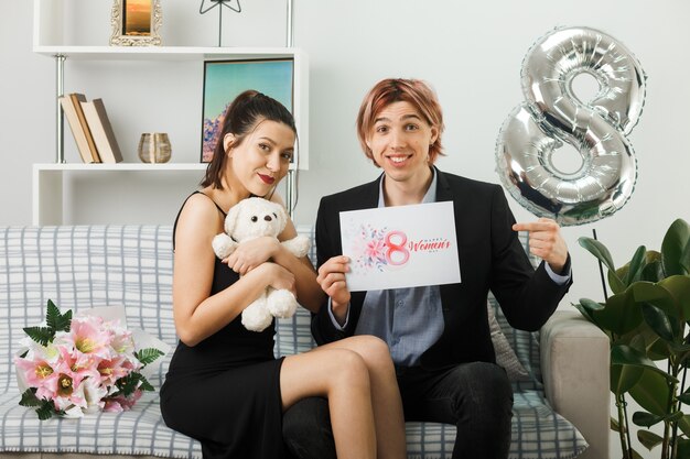 Довольная молодая пара в счастливый женский день с плюшевым мишкой и поздравительной открыткой, сидя на диване в гостиной