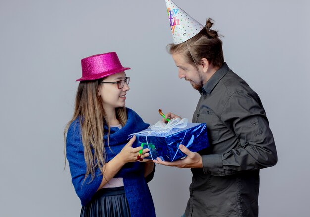 Довольная молодая пара смотрит друг на друга, держа в руках подарочную коробку, девушка в очках в розовой шляпе держит свисток, а красивый мужчина в кепке дня рождения держит свисток, изолированный на белой стене