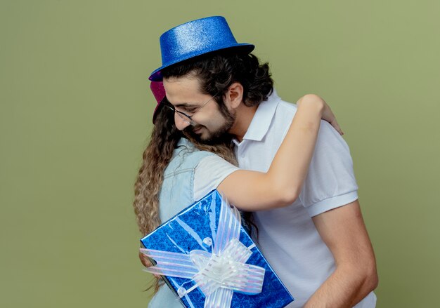 Pleased young couple wearing pink and blue hat hug each other and guy holding gift box 