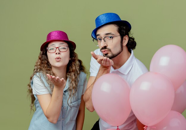 ピンクと青の帽子をかぶって風船の後ろに立ってキスジェスチャーを見せて喜んでいる若いカップル