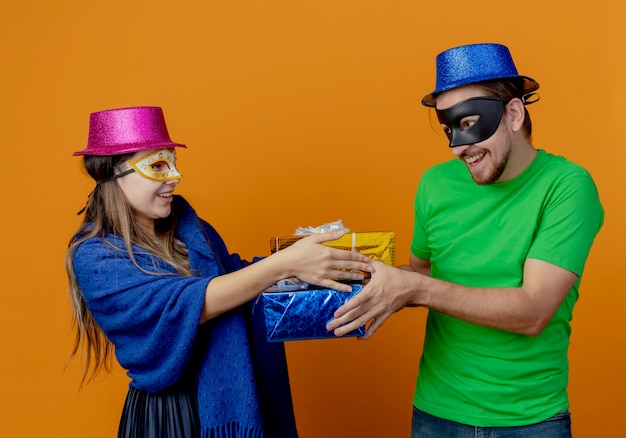 Pleased young couple wearing pink and blue hats put on masquerade eye masks looking at each other holding gift boxes isolated on orange wall