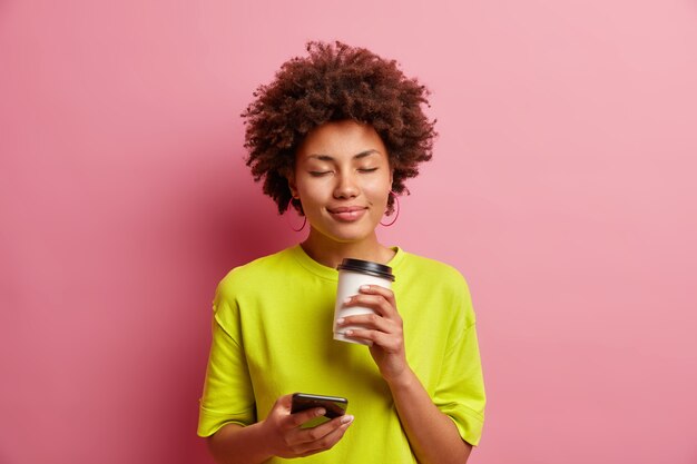 Pleased young curly Afro American woman closes eyes from smelling pleasant coffee aroma has relaxed face uses smartphone for chatting online dreessed in casual wear isolated over pink wall