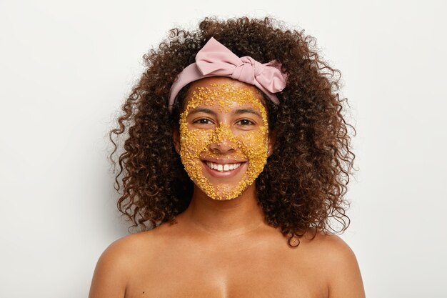 Довольная молодая темнокожая модель использует удивительный ингредиент для здоровья кожи, имеет желтые гранулы соли на лице, использует все эффективные способы быть красивой и молодой, выводит токсины, проходит гигиенические процедуры.