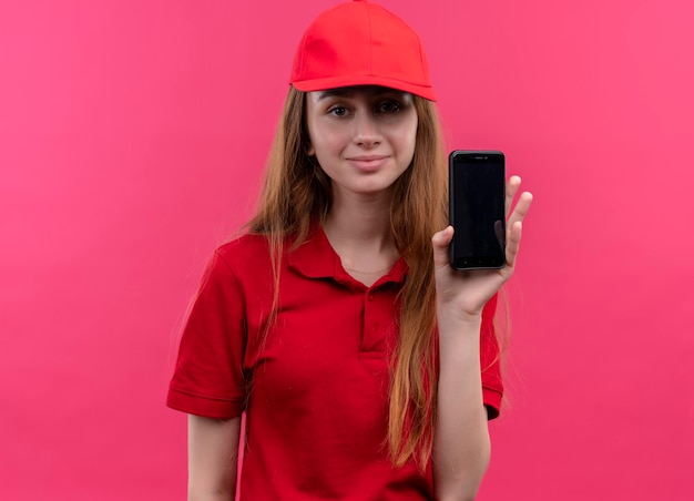 Pleased young delivery girl in red uniform holding mobile phone on isolated pink space