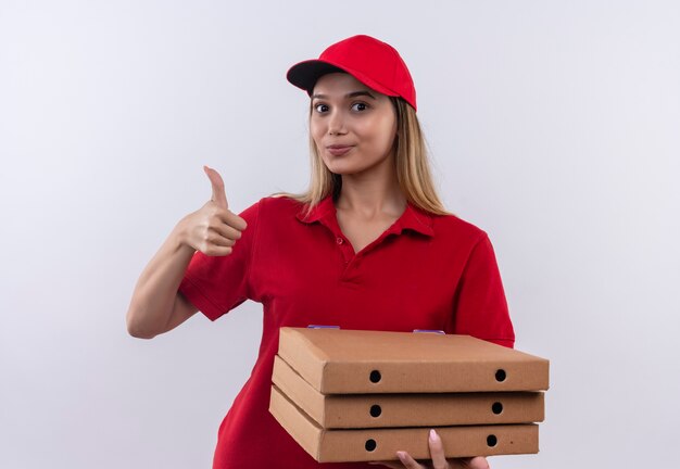 Pleased young delivery girl wearing red uniform and cap holding pizza boxes her thumb up  isolated on white wall