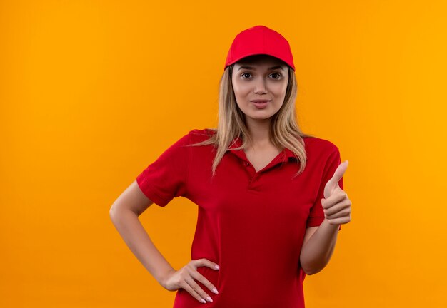 Довольная молодая доставщица в красной униформе и кепке, положив руку на бедро, подняв палец вверх, изолирована на оранжевом фоне