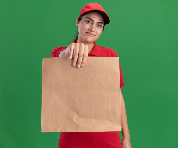 Pleased young delivery girl wearing uniform and cap holding out paper food package at front isolated on green wall