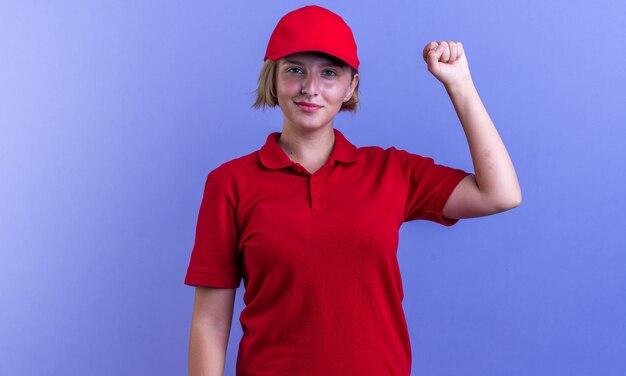 Довольная молодая доставщица в униформе и кепке показывает жест `` Да '' на синей стене