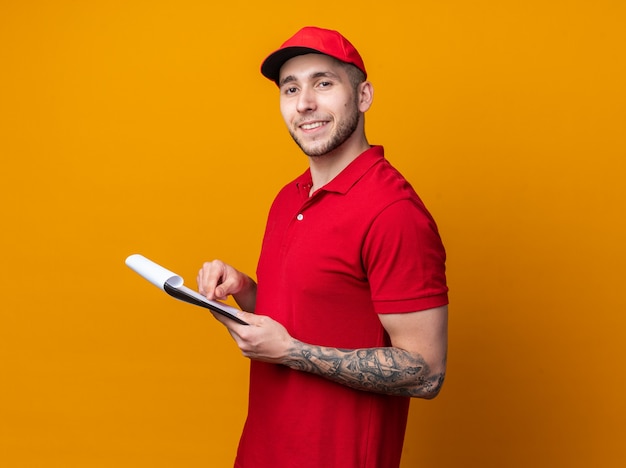 Pleased young delivery guy wearing uniform with cap holding box with clipboard 