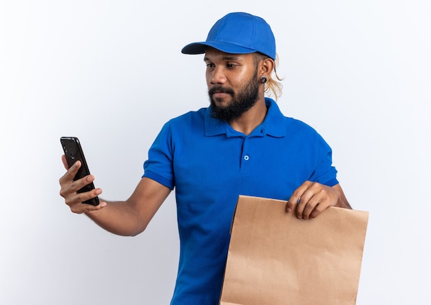 Compiaciuto giovane fattorino che tiene in mano il pacchetto alimentare e guarda il telefono isolato sul muro bianco con spazio di copia