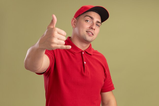 Pleased young delivery man wearing uniform and cap showing phone call gesture isolated on olive green wall