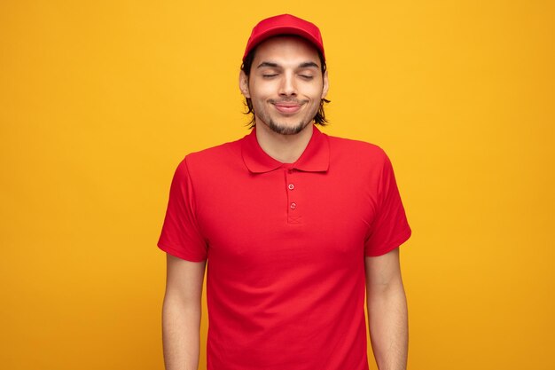 pleased young delivery man wearing uniform and cap smiling with eyes closed isolated on yellow background