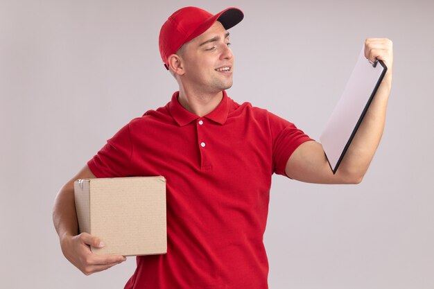 Pleased young delivery man wearing uniform with cap holding box and looking at clipboard in his hand isolated on white wall